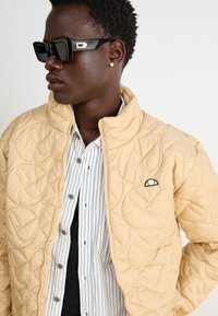 Beige vadderad jacka med hög krage, med ett cirkulärt mönster och en liten logotyp. Randig skjorta under, accessoriserad med svarta solglasögon.