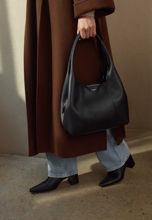 Borsa a tracolla in pelle nera con un design morbido e strutturato, dotata di chiusura con zip e un discreto dettaglio del logo. Indossata con un cappotto marrone e jeans.