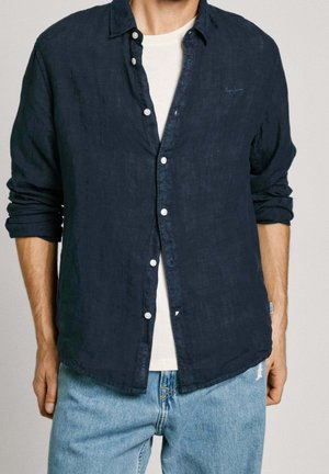 Mann trägt ein marineblaues Hemd über einem weißen T-Shirt mit hellblauen Jeans, die Ärmel hochgekrempelt, steht vor einem schlichten Hintergrund.