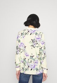 Woman with short black hair wearing a cream long-sleeve shirt with purple floral print and blue jeans, facing away against a plain background.