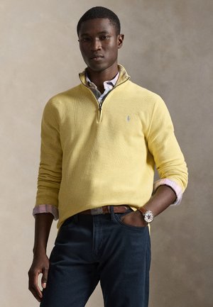 Homme portant un pull jaune à fermeture quart de zip sur une chemise rose à carreaux avec les manches retroussées, un pantalon foncé et une montre-bracelet, debout devant un fond uni.