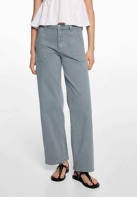 Vrouw in lichtblauwe wijde broek met voorzakken, witte mouwloze top en zwarte open teen sandalen.