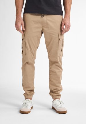 Beige cargo bukser med elastiske manchetter, flere sidelommer og en snøre i taljen. Bæret med hvide sneakers.