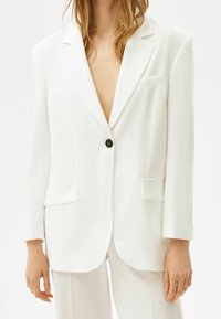 Kvinna i en överdimensionerad vit kavaj med en svart knapp och matchande vita byxor, visad från hals till mitten av låret.