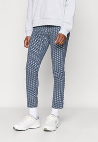 Zilās rakstainās bikses ar ģeometrisku dizainu, piegu�ļošs modelis, kombinētas ar baltām kedām un pelēku sporta krekli.