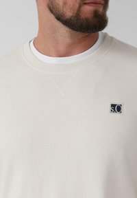 Sweat-shirt beige clair avec un col rond et une texture subtile. Logos en patch marine sur le côté gauche, superposé sur un T-shirt blanc.