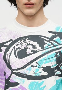 Mladá osoba v bielom tričku s okrúhlym výstrihom, ktoré má abstraktný fialovo-čierno-tyrkysový motív glóbusu a dynamického oblúka.