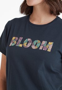 Navy blue shirt with "BLOOM" in large letters filled with colorful flower patterns worn by woman with shoulder-length brown hair.