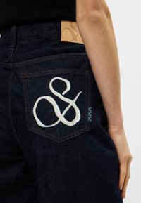 Σκούρα τζιν παντελόνια διαθέτουν ένα μεγάλο λευκό "&" κεντημένο στην πίσω τσέπη, με ελαφριά μπλε ραφή και δερμάτινη φάσα στη μέση σε απόχρωση καφέ.