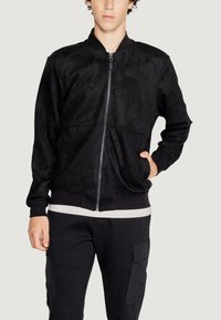 Giacca bomber nera in ecopelle, con zip frontale, polsini a coste e texture liscia. Abbinata a una maglietta bianca.