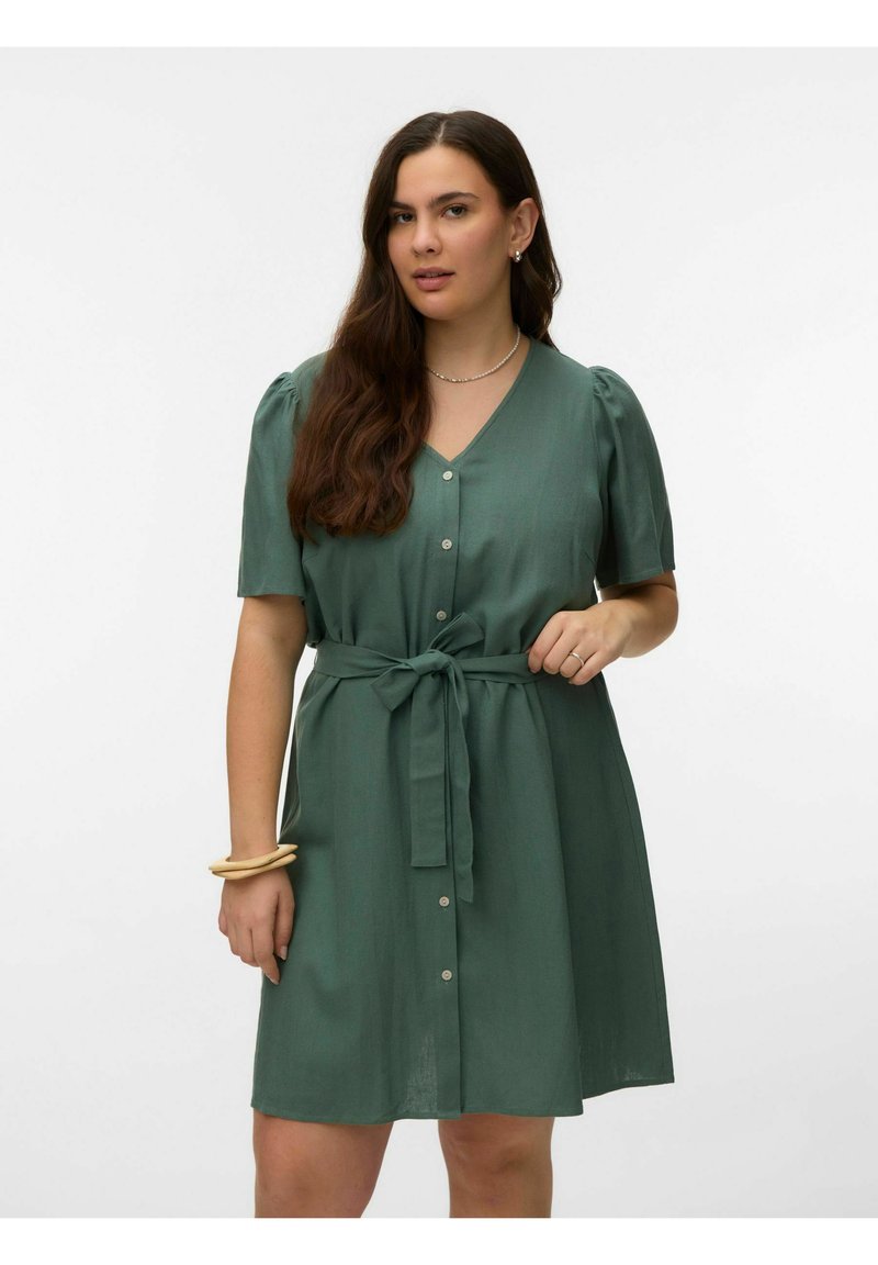 Femme portant une robe verte à manches courtes avec des boutons sur le devant, une ceinture nouée à la taille, un collier, des boucles d’oreilles et des bracelets sur un fond clair.