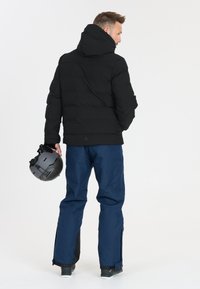 Schwarze Daunenjacke mit Kapuze, kombiniert mit marineblauen Skihosen. Die Jacke hat ein gestepptes Design, und die Hosen verfügen über Reißverschlüsse an den Knöcheln.