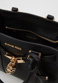 Borsa a mano in pelle nera con texture a granuli, hardware dorato e un'etichetta con logo distintivo. Presenta un vano interno con zip.