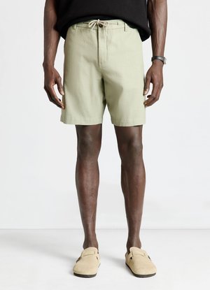Mann trägt hellbeige Shorts mit Kordelzug, schwarzes Top, beige Slip-On-Schuhe und eine schwarze Armbanduhr und steht auf einem weißen Boden.