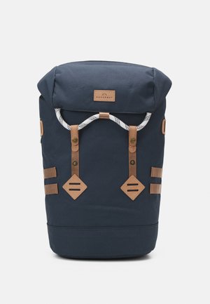 Navy blue backpack with a roll-top design, leather accents, metal hardware, and a white drawstring closure. Features two side straps and diamond patches.