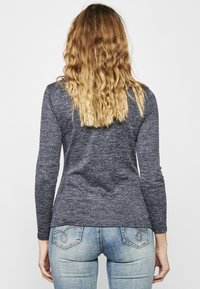 Ilga rankovė, tamsiai mėlyna viršutinė dalis, pagaminta iš tekstūruoto audinio, turi prigludusį siluetą ir šiek tiek išlenktą apačią, derinama su šviesiai mėlynomis džinsinėmis kelnėmis su galiniais kišenėmis.