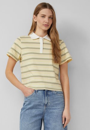 Kobieta w żółtej polo w paski z krótkim rękawem i białym kołnierzykiem oraz niebieskie dżinsy, stoi z jedną ręką w kieszeni.