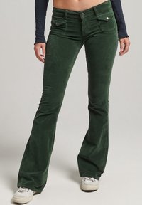 Pantalons évasés en velours côtelé vert avec poches avant, une texture lisse et une taille à bouton. Associés à des baskets blanches.