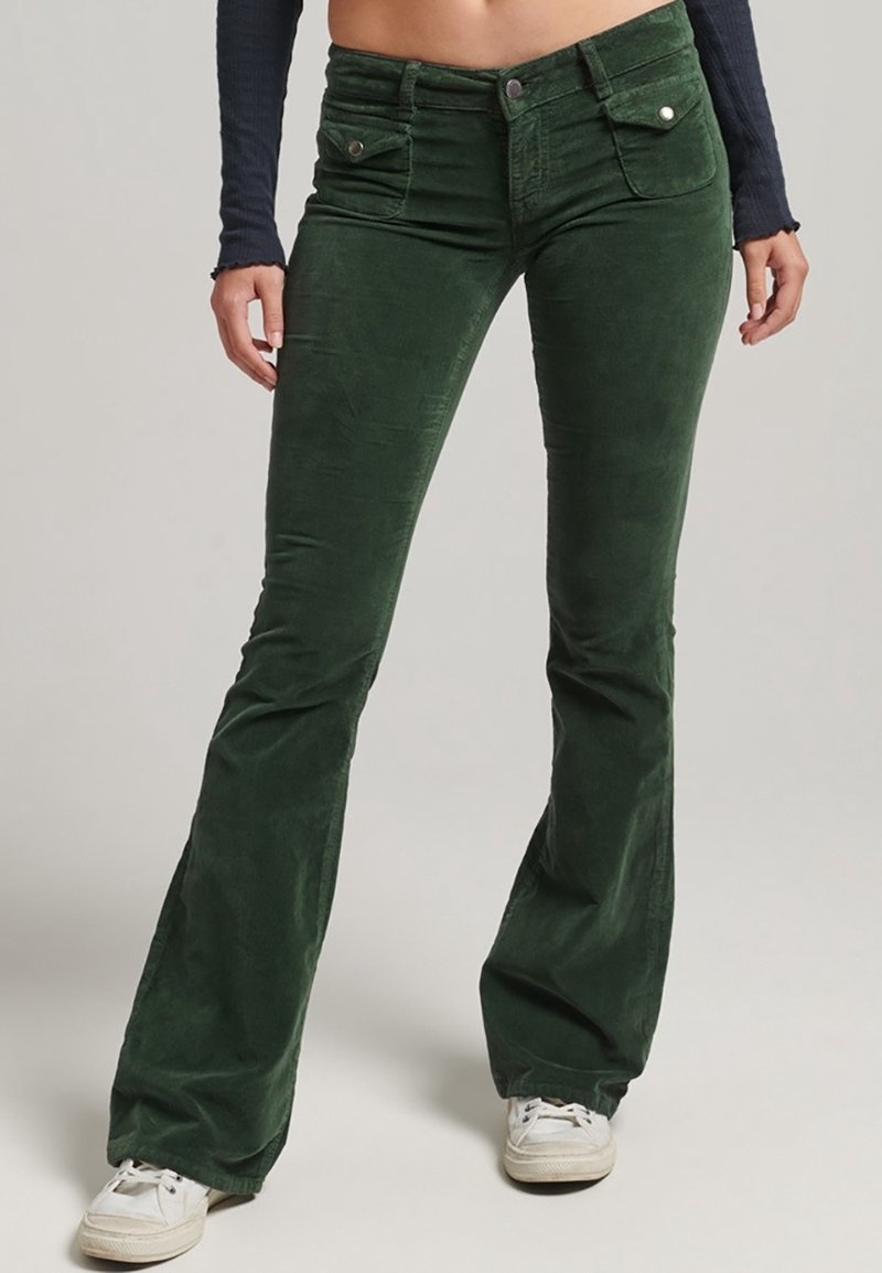 Pantalons évasés en velours côtelé vert avec poches avant, une texture lisse et une taille à bouton. Associés à des baskets blanches.