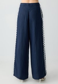 Pantalon large marine en tissu léger, avec des bandes latérales blanches à volants. Coupe ample avec une texture lisse, coupé au niveau de la cheville.