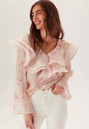 Donna sorridente con lunghi capelli castani indossa una camicetta trasparente, arricciata e floreale e pantaloni bianchi, posando su uno sfondo chiaro e uniforme.