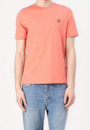 Mand iført koralfarvet T-shirt med korte ærmer og lille gult fuglelogo på brystet samt lyseblå jeans, stående mod en hvid baggrund.