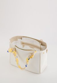 Witte canvas handtas met een gestructureerd ontwerp, beige accenten, gouden hardware en een meerkleurige bloemenpatroon sjaal die aan de handgreep is geknoopt.