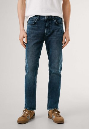 Donkerblauwe denim jeans met een rechte pasvorm, vijf zakken en een subtiele vervaagde afwerking. Gecombineerd met bruine sneakers met blauwe accenten.