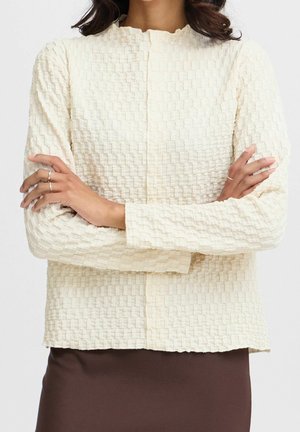 Femme aux bras croisés portant un haut crème texturé à manches longues et une jupe marron foncé, montrant de délicates bagues aux doigts.