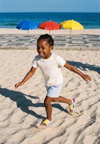 Kleines Kind in weißem Hemd und hellblauer Shorts läuft fröhlich am Sandstrand mit bunten Sonnenschirmen und Ozean im Hintergrund.