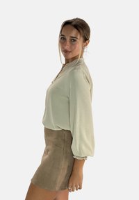 Licht beige blouse met een gestructureerde afwerking, voorzien van een opstaande kraag en lange pofmouwen. Gepaard met een korte, tan suède rok.