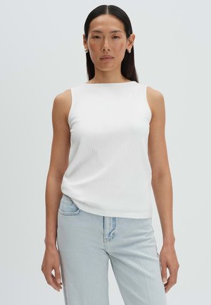 Donna con capelli lunghi e scuri che indossa una canottiera bianca a coste e jeans azzurri chiari, in piedi davanti a uno sfondo grigio chiaro uniforme.