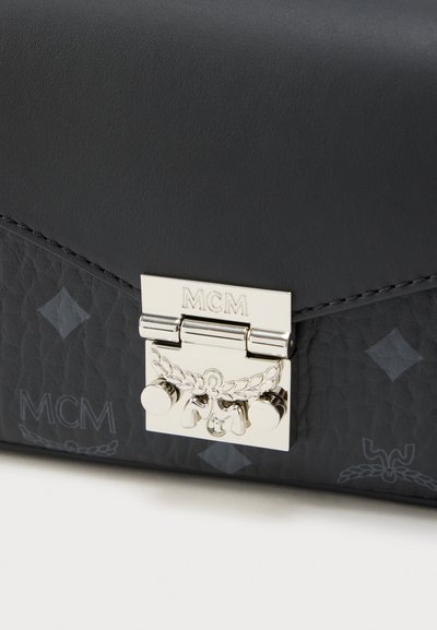 Γωνία μαύρης δερμάτινης τσάντας με υφή, με ασημένιο μεταλλικό κούμπωμα κλειδώματος χαραγμένο με το «MCM» και σχέδιο από στέφανο δάφνης, καθώς και διακριτικές εκτυπώσεις λογότυπου MCM.