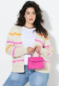 Strik cardigan i creme med lyserøde og orange mønstre, parret med en lyserød taske med krokodillepræg. Modellen har en hvid top og blå jeans på.
