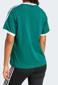 Persona vistiendo una camiseta deportiva de manga corta en color teal con ribete blanco y leggings negros con rayas blancas, mostrada desde atrás.