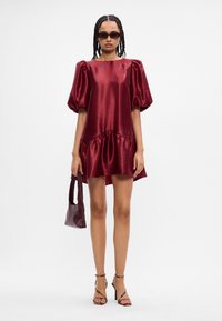 Βουργουντί σατέν φόρεμα με φουσκωτά μανίκια, ριχτό τελείωμα και χαλαρή εφαρμογή. Το μοντέλο έχει αξεσουάρ ένα μικρό σκούρο τσαντάκι και ψηλοτάκουνα σανδάλια.