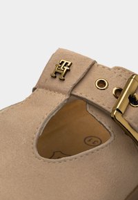 Μπεζ σουέτ παπούτσι Mary Jane με χρυσή αγκράφα και λογότυπο TH, με καρδιοειδές άνοιγμα κοντά στο λουράκι.