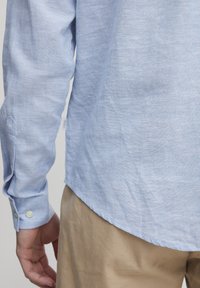 Mies, joka on pukeutunut vaaleansiniseen pitkähihaiseen paitaan, jossa on napit hihansuissa, ja vaaleisiin housuihin, kuvattuna selkäpuolelta vyötärön tasolla.