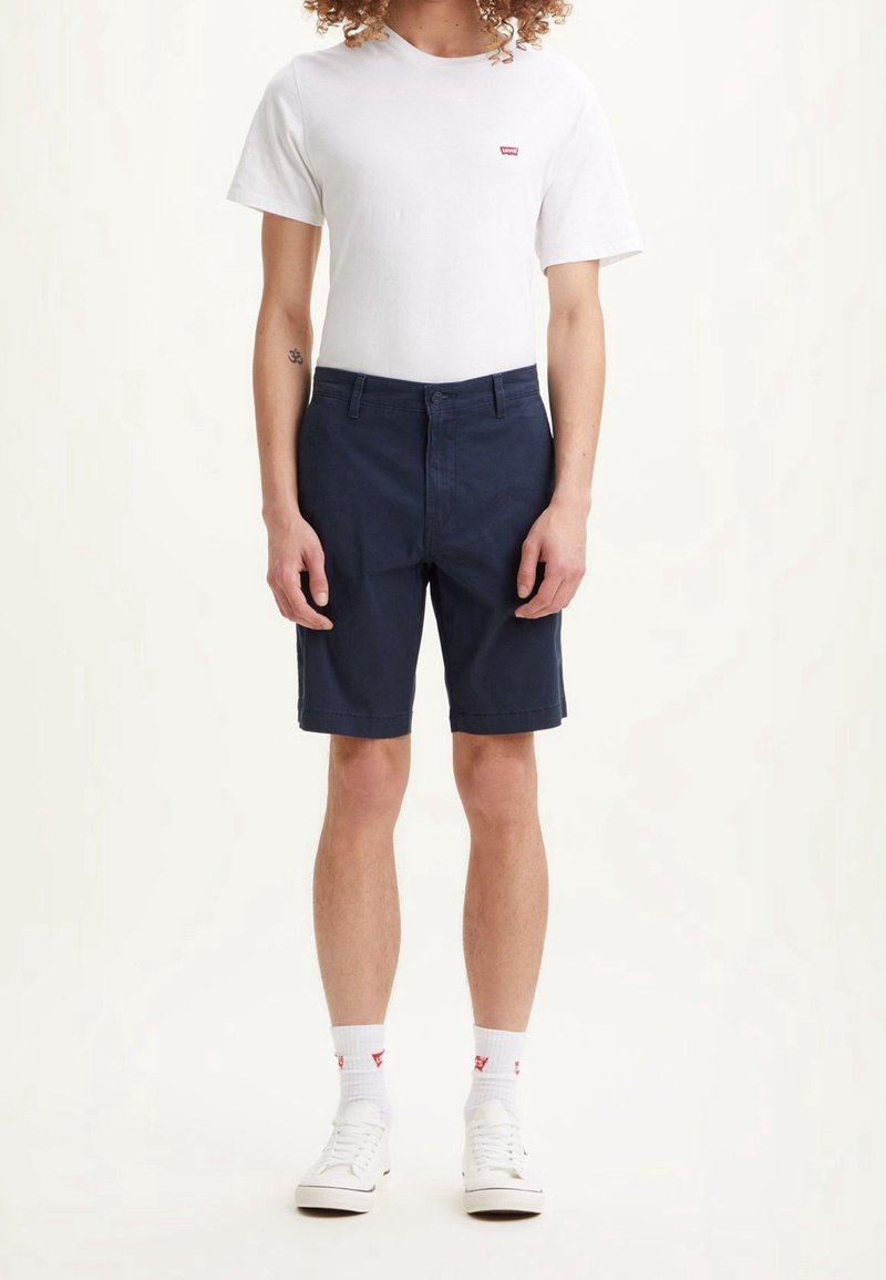Personne portant un t-shirt blanc Levi's, un short bleu marine, des chaussettes blanches avec un logo rouge, et des baskets blanches, debout devant un fond uni.