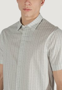Camicetta a maniche corte in grigio chiaro con un motivo geometrico ripetuto. Presenta un colletto button-down e una texture liscia e morbida.
