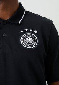 Černé polo tričko s bílým lemem má na levém prsu logo Deutsche Fussball-Bund a čtyři hvězdy nad ním, nošené osobou.