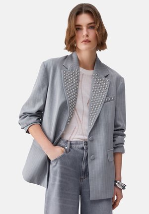 Donna che indossa un blazer grigio a righe sottili con borchie argento sulle rever, una camicia bianca strutturata e jeans grigi a vita alta con la mano in tasca.