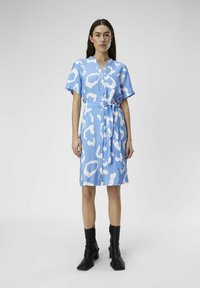 Hellblaues Hemdkleid mit weißem abstraktem Leopardenmuster, kurzen Ärmeln, einem taillierten Gürtel und einem leicht strukturierten Stoff. Schwarze Knöchelstiefel.