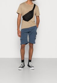 Beige katoenen T-shirt, blauwe cargoshort met zijzakken, zwarte crossbodytas en zwarte hoge sneakers. Effen achtergrond.