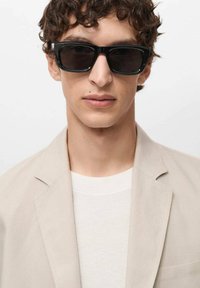 Black rectangular sunglasses with a thick frame worn with a light beige blazer over a white textured knit sweater. Smooth surfaces and clean lines.