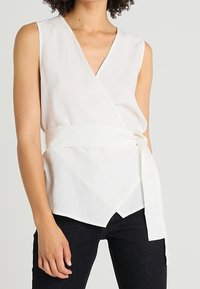 Top bianco senza maniche a wrap con scollo a V profondo, cintura auto-tie e design strutturato, caratterizzato da un tessuto liscio e orlo dritto.