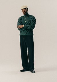 Άνδρας με μπεζ καπέλο, χρυσά γυαλιά, πράσινο πουλόβερ με λαιμόκοψη, μαύρο παντελόνι, μαύρα παπούτσια και χρυσά κοσμήματα, στέκεται με τα χέρια σταυρωμένα.