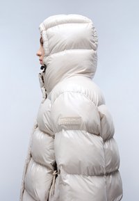 Giacca puffer beige con finitura lucida, cappuccio e design a coste. Presenta un patch con logo sulla manica sinistra e dettagli arricciati all'orlo.