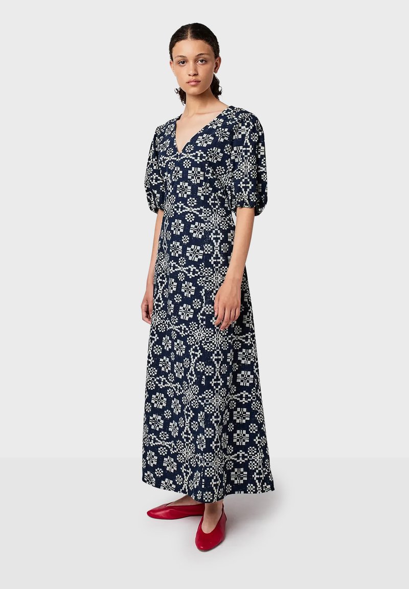 Žena stojí oblečená v dlouhé šaty s geometrickým vzorem v námořnické modré a bílé barvě, s nafouknutými rukávy a červenými plochými botami, na jemném světlém pozadí.