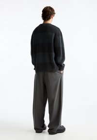 Jeune personne vue de dos, portant un pull en maille à rayures sombres, un pantalon large gris et des chaussures noires épaisses, les mains dans les poches.